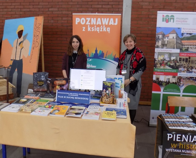 Panie stoją obok stolików, na których ułożone są książki i czasopisma. W tle baner PBW w Krakowie.