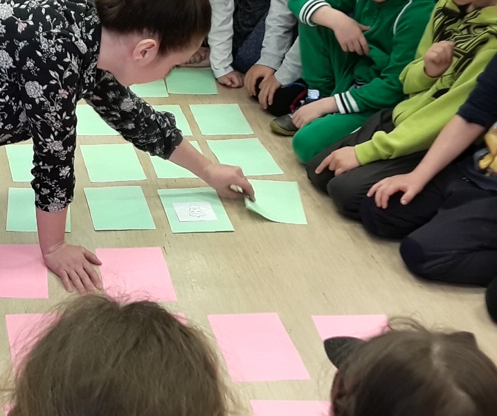 Na dywanie rozłożone kartki zielone i różowe. Pani ręką odkrywa zieloną kartkę i wyjaśnia dzieciom zasady gry.