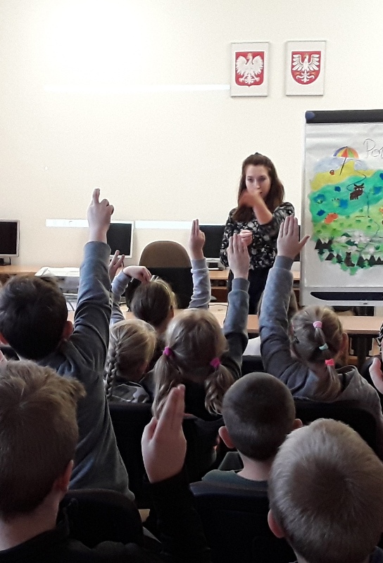 Na zdjęciu dzieci stoją tyłem do obiektywu, podnoszą rękę, zgłaszając gotowość do odpowiedzi. Przed dziećmi stoi pani, która wskazuje dłonią w kierunku ucznia. Z tyłu po prawej stronie narysowana geograficzna mapa Polski.