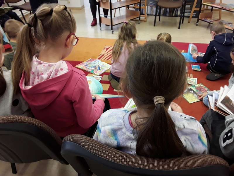 Dwie dziewczynki siedzą tyłem do obiektywu i oglądają książeczki z baśniami. Na drugim planie na dywanie siedzą dzieci zwrócone tyłem do obiektywu, które przeglądają książeczki. Na dalszym planie widać regały i krzesła.