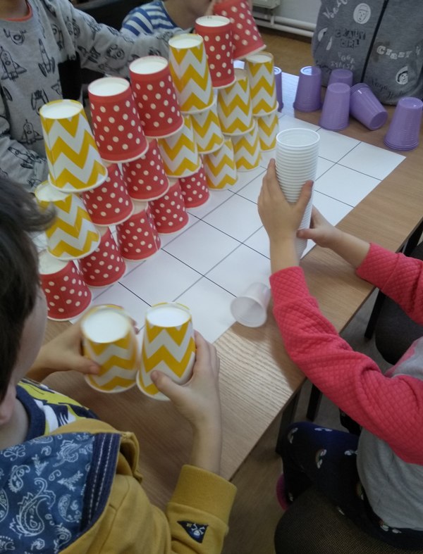 Na stoliku znajduje się biała kartka papieru z narysowaną szachownicą, na której stoi wieża zbudowana z plastikowych kubeczków biało żółtych i biało czerwonych. Obok stoją kubki koloru fioletowego. Na zdjęciu widać ręce dzieci, które układają wieżę.