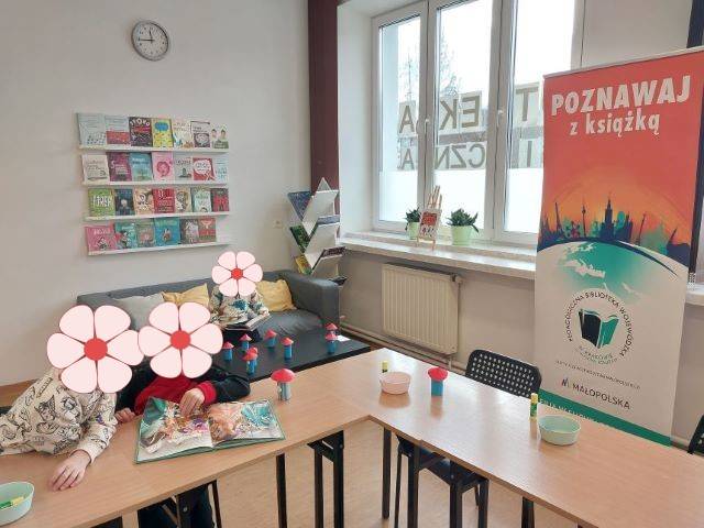 Dzieci siedzą przy stolikach i oglądają książki.